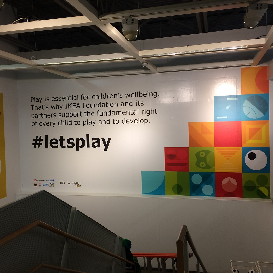 A printed wall mural installed above a staircase at IKEA reading '#letsplay', paired with IKEA Foundation partner logos and a colourful geometric pattern block