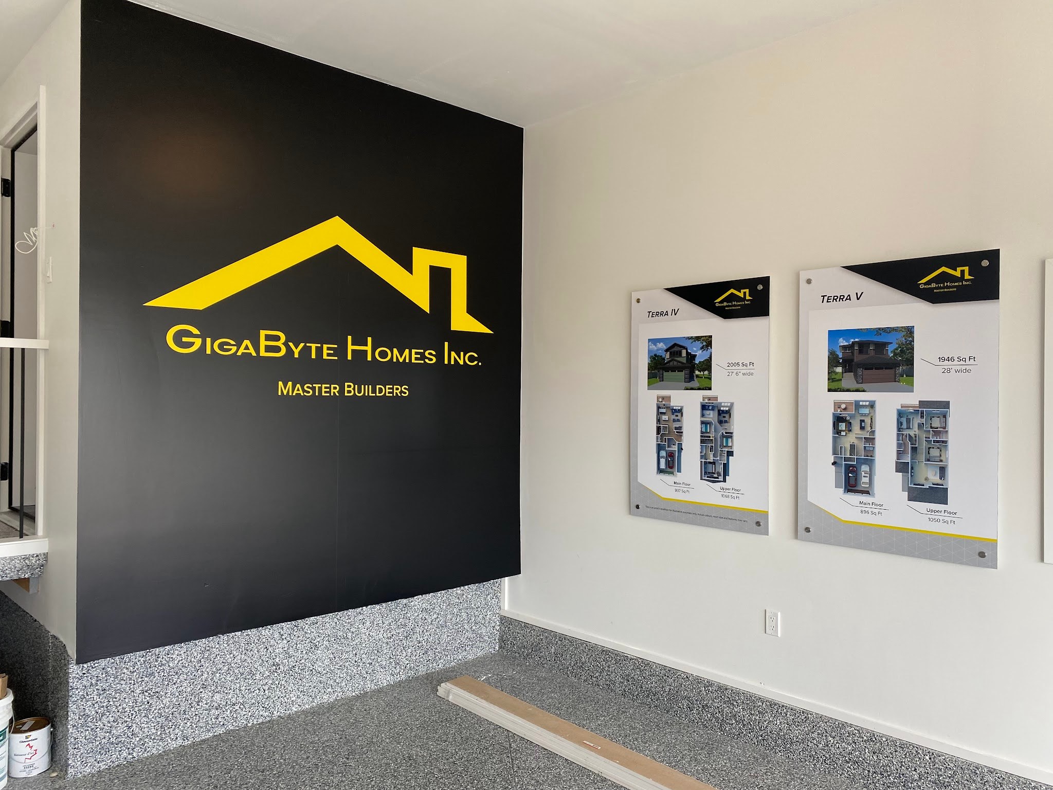 Printed architectural wall graphic on a black feature wall in a builder's show home sales space, carrying the GigaByte Homes roofline logo beside a display of framed floor plans