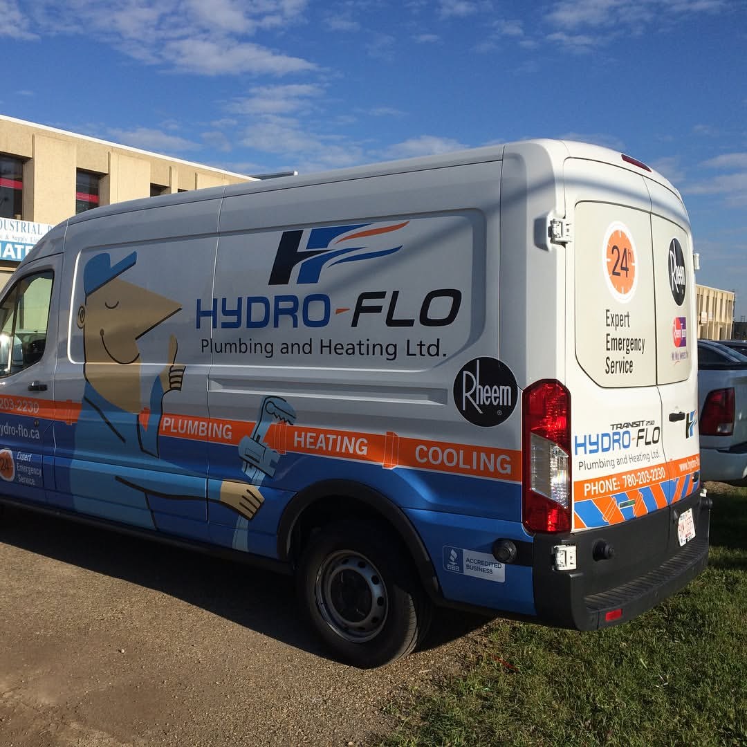 Rear three-quarter view of a Hydro-Flo Plumbing and Heating Ford Transit in a full wrap: cartoon plumber on the side, an orange band reading plumbing, heating, cooling, and the 24-hour emergency badge on the rear doors
