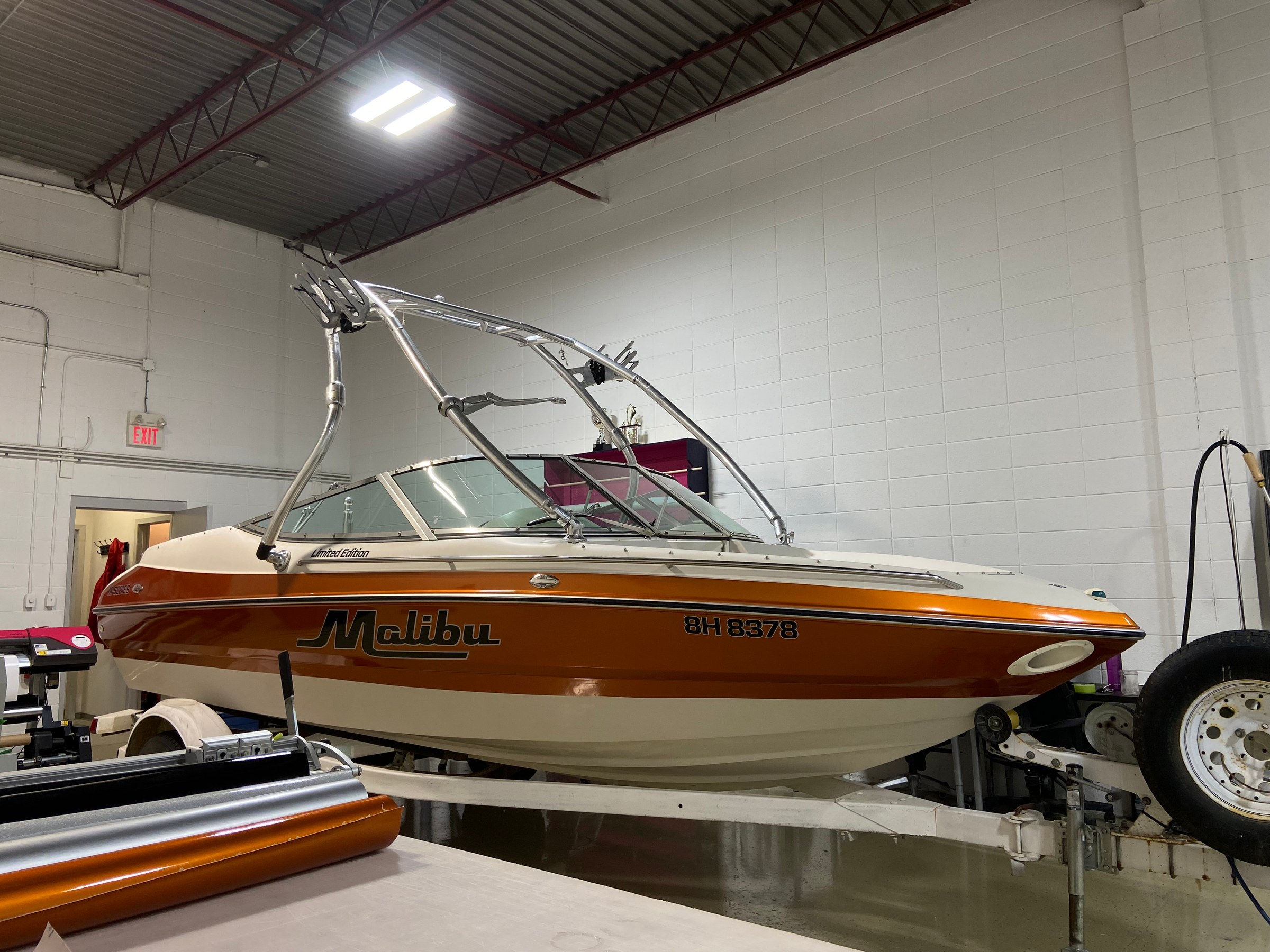 Malibu M-Series wakeboat in an orange metallic wrap with printed Malibu graphics along the hull, on its trailer in the shop