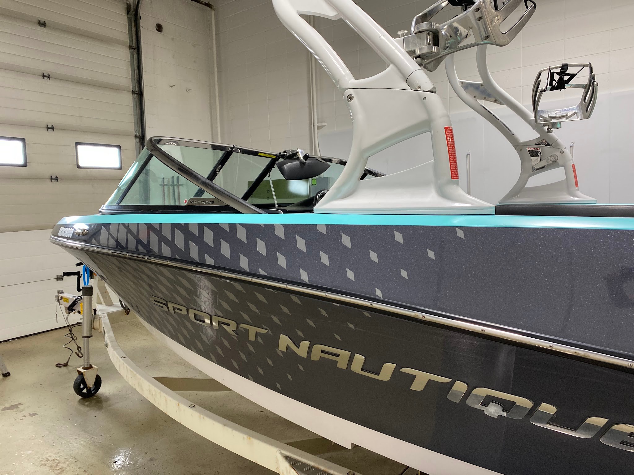 Bow and hull side of a Sport Nautique wakeboat wrapped in satin Key West teal with a dark grey lower hull, on its trailer in the shop