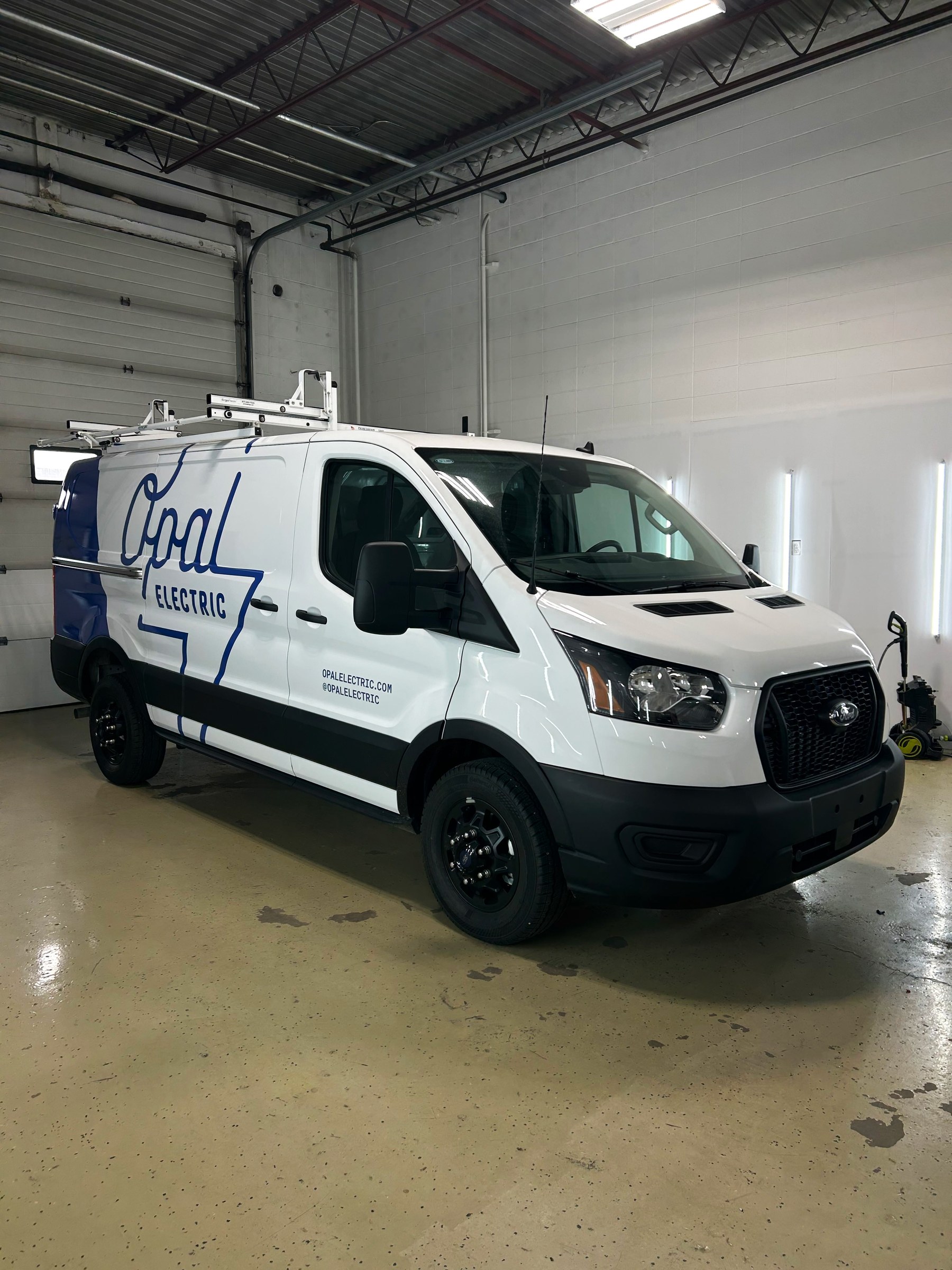 A Coal Electric Ford Transit service van in a white and blue wrap with a roof rack, photographed front three quarter in the shop